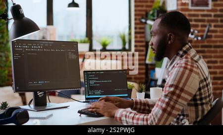 Young man seen programming software code on his laptop in a sleek home office. Workspace with dual monitors helps with software engineering, web development and coding productivity. Camera B. Stock Photo