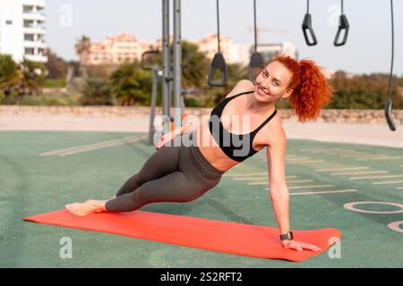 Happy sporty woman practicing side plank position on exercise mat at outdoor gym. Portrait of smiling young female in activewear is working out at city park during daytime.  Stock Photo