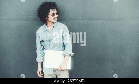 Young biracial man with curly hair wearing sunglasses and a casual denim shirt stands against a textured green wall, holding a closed laptop and looki Stock Photo