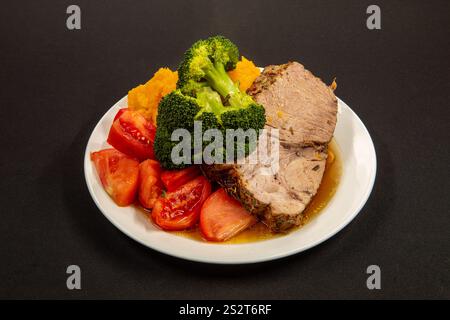 Thick, juicy slce of roasted pork, accompanied by ripe tomatoes and green broccoli, arranged on a simple white dish. Side view. Black background.  Res Stock Photo
