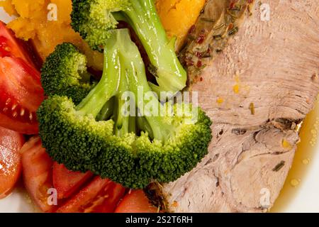 A close-up view of a thick slice of roasted pork, surrounded by fresh tomatoes and broccoli, served on a clean white plate.  Restaurant Menu. Stock Photo