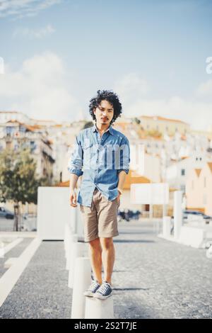 A young biracial man with curly black hair stands confidently on a concrete pillar in an urban outdoor setting. He wears a denim shirt, khaki shorts, Stock Photo
