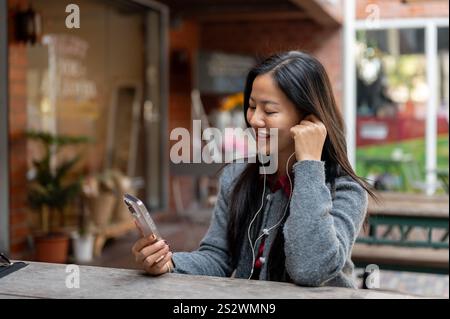 A positive Asian woman enjoys listening to music on her earphones and using her smartphone while sitting outdoor in the city. people, technology, life Stock Photo