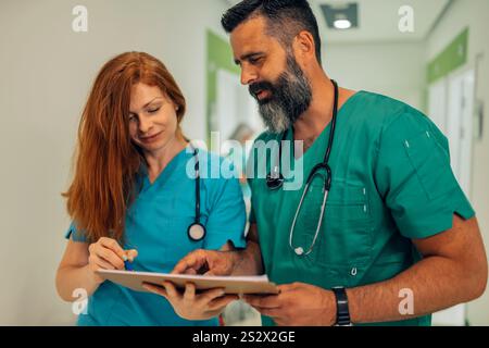 Two medical professionals, a female physician with red hair and a male doctor with a beard, are working together to review details on a clipboard in a Stock Photo