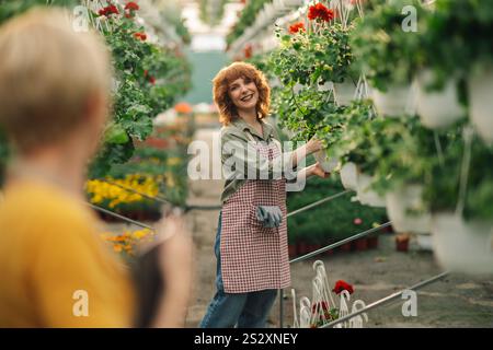 Portrait of young cheerful ginger florist standing near hanging pots at greenhouse and working with flowers while smiling at camera. Young botany expe Stock Photo