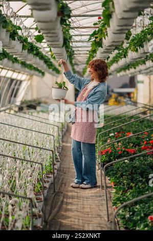 Full length portrait of smiling female ginger botanist standing at hothouse with pot of flower sapling surrounded by flowers and plants. Young redhead Stock Photo