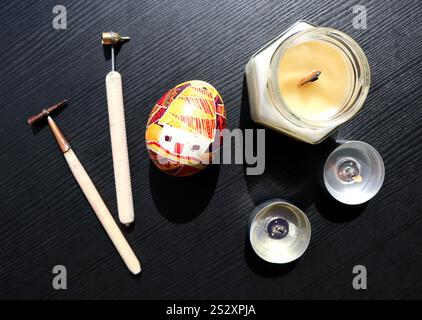 Easter egg. A house is painted on the egg, geometric ornaments of different colors. Nearby are various candles, tools for coloring eggs. Isolated on a black wooden background. Photo. Easter tradition. Stock Photo