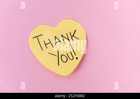 A single yellow, heart-shaped Post-It note with the handwritten message "Thank You," placed on a pastel pink background, expressing gratitude and appr Stock Photo