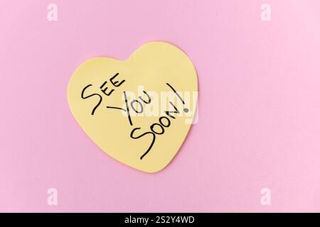 A single yellow, heart-shaped Post-It note with the handwritten message "See You Soon," placed on a pastel pink background, conveying a sweet farewell Stock Photo