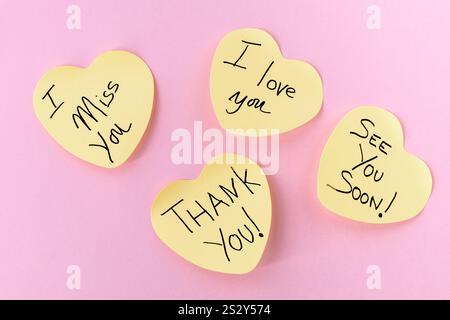 Four yellow, heart-shaped Post-It notes with handwritten messages: "I love you," "I miss you," "Thank you," and "See you soon," arranged on a pastel p Stock Photo