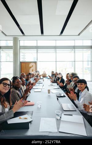 Diverse group of people clapping and looking at camera during a business meeting in a bright, modern office setting. Stock Photo