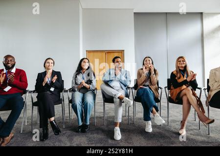 Attentive group of diverse professionals attending a corporate workshop, sitting in a row. They are smiling and applauding the speaker or presentation Stock Photo