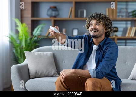 Joyful man turns on the air conditioner, sits with the remote control in his hands from the smart home system, on the sofa in the living room at home. Stock Photo