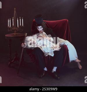 Man in black suit, vampire, holds woman dressed in white on his lap, gazing intently, next to vintage candelabra. Stock Photo