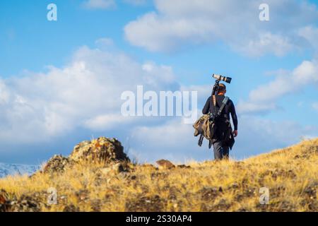 Outdoor scene featuring a photographer carrying camera equipment walking over grassy plain. Bright clear skies and natural surroundings evoke freedom exploration and creativity in untamed environment Stock Photo