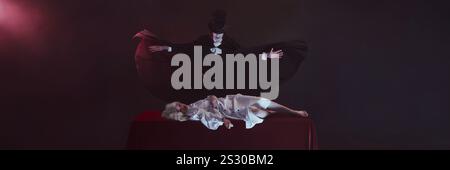 Banner. Vampire stands with arms spread wide, black cloak flowing, over table where woman dressed in white lies still, creating dramatic and eerie Stock Photo
