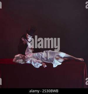 Vampire leans over woman dressed in white lying on table with red cloth, holding their wrist as if about to bite, with intense and haunting Stock Photo