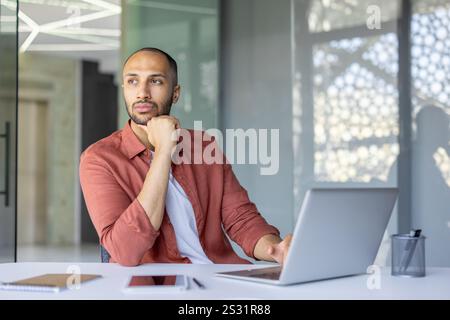 Serious thinking businessman at workplace inside office working with laptop, man in shirt pondering over solution of current financial problems tasks. Stock Photo