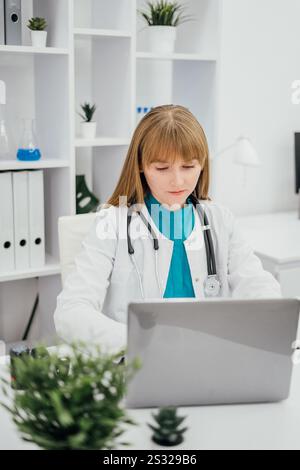 A female doctor in a white coat with a stethoscope around her neck is working on a laptop in a modern, clean office. The background features shelves Stock Photo