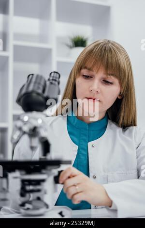 A dedicated scientist in a white lab coat and blue uniform is carefully adjusting a microscope to observe samples in detail. The modern laboratory Stock Photo
