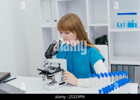 A young scientist wearing a blue uniform is focused on observing samples through a microscope in a modern laboratory. The setting includes shelves Stock Photo