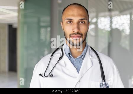 Close-up portrait of a doctor in a white medical coat, a man looks seriously and focused at the camera, the doctor works inside the clinic office at his workplace. Stock Photo