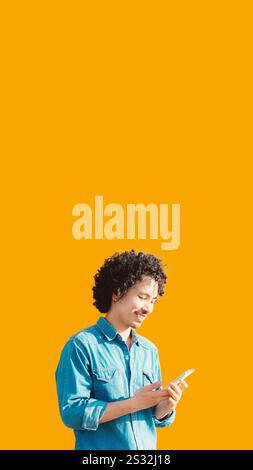 Young man with curly hair in a blue denim shirt smiling while using a smartphone against a vibrant yellow background. Ample copy space area above for Stock Photo