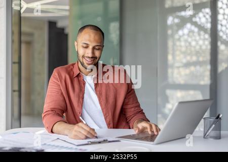 Portrait of a young successful financier, a man behind paperwork, filling out tax forms, and investment reports. Businessman at workplace with documents and laptop. Stock Photo