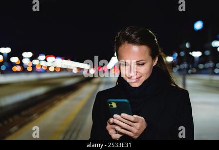 Woman using her smartphone on railway station platform at night. Person waiting for a train Stock Photo