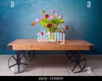 Tulips bouquet pink and red dying in glass vase, falling flowers and leaves, standing on wooden table with metal black wheels, tiled floor Stock Photo