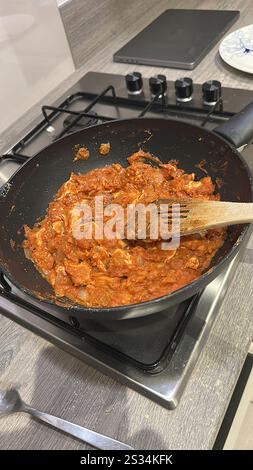 A delicious homemade chicken Thai satay curry being cooked in a black wok pan on a stove top with a wooden spoon stirring the rich tomato-based sauce Stock Photo