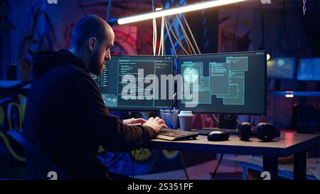 Hacker in underground warehouse writing lines of code on computer screen to make malware that get past security systems. Man in hidden HQ building scripts that can hack devices, dolly in shot Stock Photo