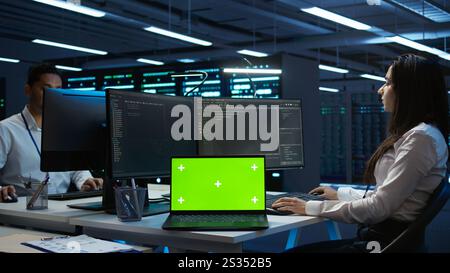 Programmers in data center manage and maintain databases using chroma key notebook, ensuring data integrity and security. Experts updating server room systems using isolated screen laptop, camera B Stock Photo
