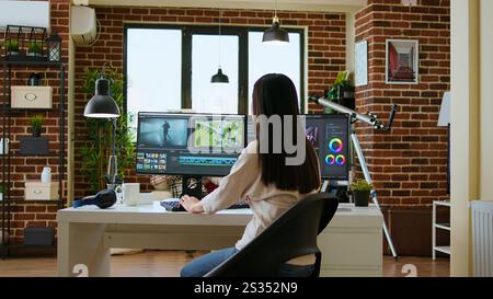 Asian woman edits videos in a creative software at her home office. Filmmaker using dual monitors and color wheel to work on footage in post production, highlighting remote work lifestyle. Camera A. Stock Photo