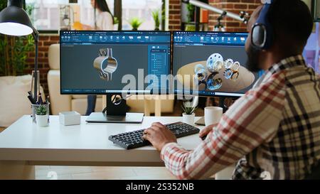 African american industrial designer works on CAD software and listens to music, multitasking at home. Engineer specialist uses digital tools and 3D modeling for industrial projects. Camera A. Stock Photo