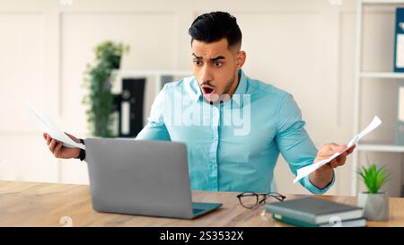 Shocked Arab businessman with documents looking at laptop screen, shouting WTF, making mistake in project at office Stock Photo