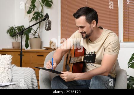 Young happy male composer with guitar writing notes on armchair at home Stock Photo