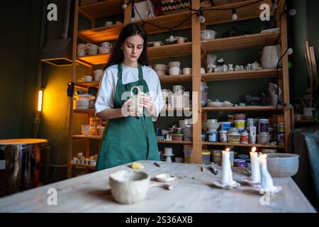 Female potter check molded clay mug for strength, touching handle in workshop. Craftsmanship skills Stock Photo