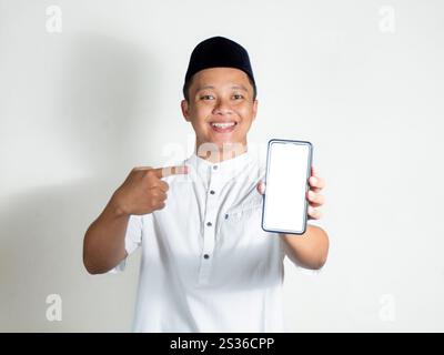 Portrait of Moslem Asian man smiling and showing smartphone blank screen. Moslem wearing white shirt and black cap Stock Photo