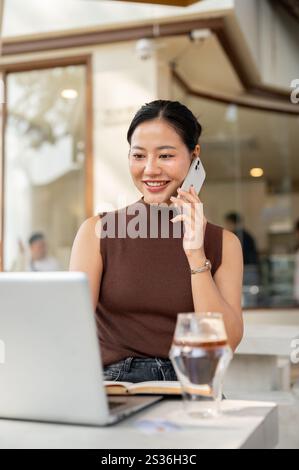 A beautiful, confident Asian woman talks in the phone while working remotely from a cafe in the city. freelancer, businesswoman Stock Photo