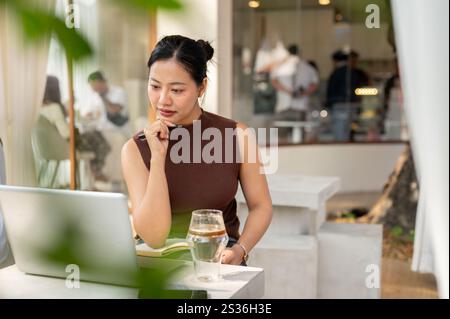 A beautiful, focused Asian woman reads something on her laptop screen, working remotely from a cafe in the city. people and lifestyle concepts Stock Photo