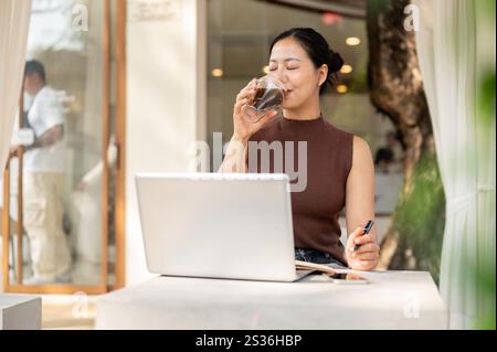 A satisfied, relaxed Asian woman enjoys a glass of coffee while working remotely in a cafe, seated in front of her laptop. people and lifestyles Stock Photo