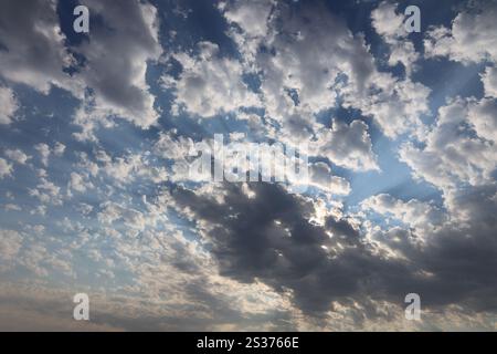 rays of setting sun stream through gaps in light puffy clouds against blue sky Stock Photo