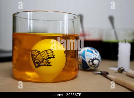 Process of drawing Easter eggs. Glass filled with yellow liquid. It contains an egg with ornamental bird. Nearby candle, painted egg, tools for drawing on eggs. Other glasses on background. Photo. Stock Photo