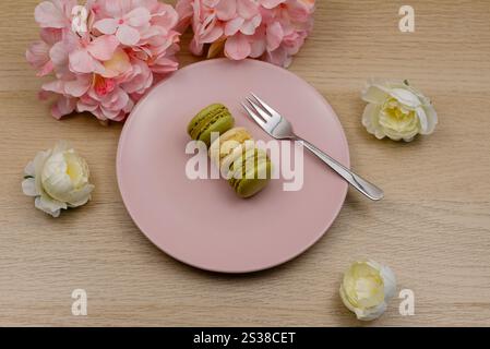 Pistachio and vanilla macarons on a pink plate on a wooden table with in the background pink and cream coloured flowers Stock Photo