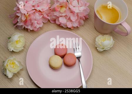 A mug of coffee with French raspberry and vanilla macarons on a wooden table with pink and cream coloured flowers. Romantic background Stock Photo