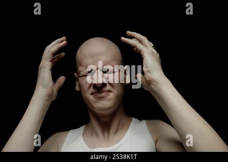 Portrait of troubled bald man with screwed up eyes experiencing intense headache raising arms in anguishing feeling while standing against black background in studio with low key light, copy space Stock Photo