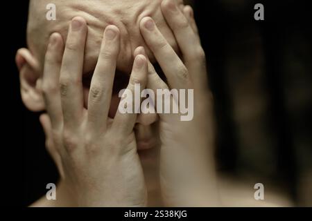 Close up on pale bald man covering face with hands suffering from acute migraine pain captured in dark studio with loy key light, copy space Stock Photo