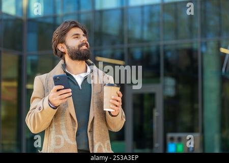 Happy Indian businessman enjoying morning coffee hot drink and using smartphone sending messages outdoors. Relaxing, taking a break. Man guy standing on urban city center street, drinking coffee to go Stock Photo
