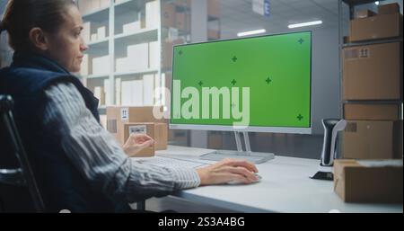 Distribution Center: Female Logistics Specialist Works on Computer in Warehouse Facility, Enters Data, Checks Inventory or Parcels Information. PC Screen Shows Green Screen Chroma Key. Dolly Shot. Stock Photo